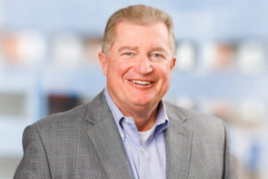 Professional headshot of Brian Snader smiling, featuring a neutral, slightly textured studio backdrop.
